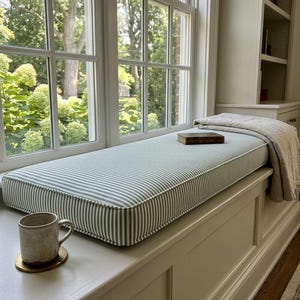May include: A rectangular window seat cushion featuring a sage green and white pinstripe pattern with white piping edges. A beige linen throw blanket is folded on one end, and a small brown hardcover book rests on the center of the cushion surface.
