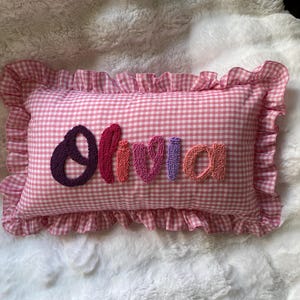 May include: A pink gingham cushion with a frilled edge. The name "Olivia" is embroidered in textured, colourful letters across the centre. A decorative item, ideal for a child's room.