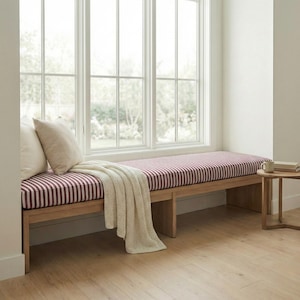 May include: A wooden bench featuring a long, rectangular cushion with a burgundy and white vertical striped pattern. Two cream-colored throw pillows are placed on the left side, and a cream-colored knit throw blanket is draped over the edge.