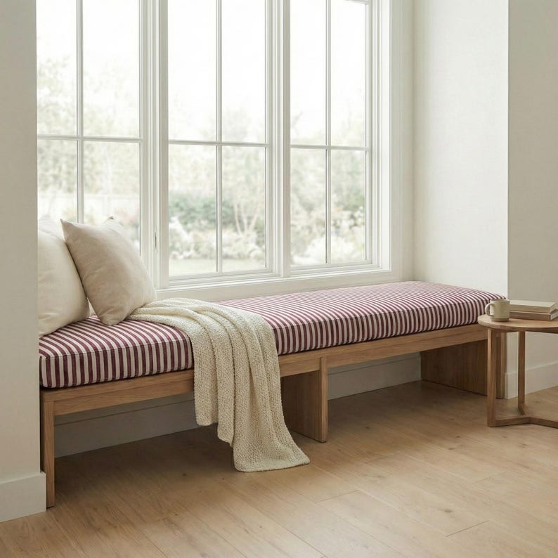 May include: A wooden bench featuring a long, rectangular cushion with a burgundy and white vertical striped pattern. Two cream-colored throw pillows are placed on the left side, and a cream-colored knit throw blanket is draped over the edge.