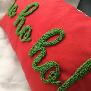 May include: A red rectangular cushion with the words "ho ho ho!" embroidered in green. The cushion has a textured appearance and is likely made of fabric. The design is festive and suitable for Christmas.