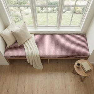 May include: A wooden window bench featuring a long cushion with a red and white vertical striped pattern. Two solid cream-colored square throw pillows and a cream-colored textured throw blanket are placed on the bench. A small round wooden side table.