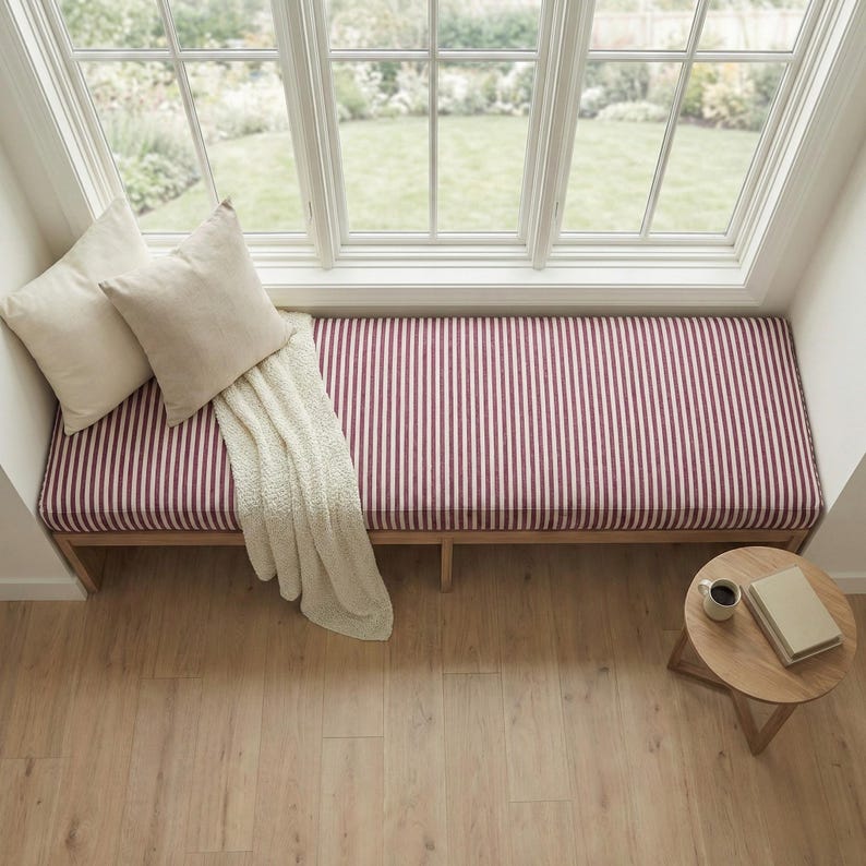 May include: A wooden window bench featuring a long cushion with a red and white vertical striped pattern. Two solid cream-colored square throw pillows and a cream-colored textured throw blanket are placed on the bench. A small round wooden side table.
