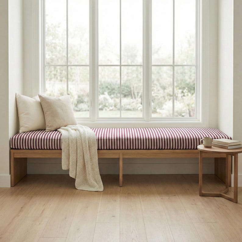 May include: A wooden bench featuring a long cushion with a burgundy and white vertical stripe pattern. Two off-white square throw pillows and a cream-colored knit throw blanket are placed on the bench. A small wooden side table sits nearby.