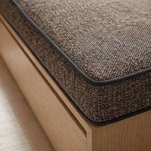 May include: A bench featuring a light-toned wooden base and a thick, rectangular seat cushion upholstered in a textured, dark brown woven fabric. The cushion is finished with a dark grey piping detail along the edge, creating a clean, defined border.