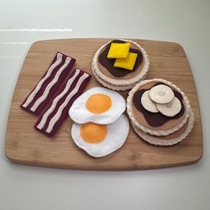 May include: A felt play food set featuring two strips of maroon bacon with white wavy lines, two white fried eggs with orange centers, a stack of pancakes topped with brown syrup and two yellow butter squares, and a stack with banana slices.