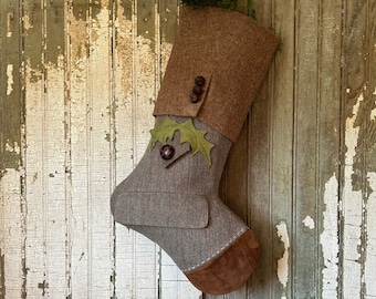 Könnte beinhalten: Ein brauner und grauer Filz-Weihnachtsstrumpf mit einem braunen Wildleder-Bund und grünen Filzblättern. Der Strumpf hängt an einer verwitterten Holzwand.