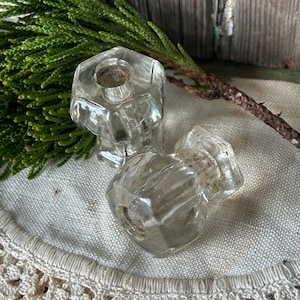 May include: Two clear, faceted glass candle holders. The candle holders have a hexagonal shape and a small opening at the top for a candle. They are resting on a cream-colored surface with a green sprig in the background.