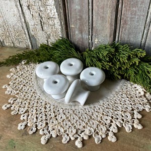 May include: A collection of white ceramic cabinet knobs with a donut-like shape, displayed on a cream-colored, crocheted doily. Green sprigs of foliage are arranged around the knobs, set against a weathered wooden background.