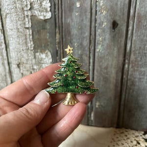 Peut inclure: Broche en forme de sapin de Noël avec une base et une étoile dorées. La broche est verte avec des accents rouges, et l'arbre a un design détaillé. La broche est tenue dans une main.