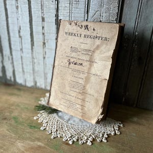 May include: An antique book titled "Weekly Register" with faded text and a worn cover. The book is propped up on a white lace doily and a wooden surface. The book's cover includes the text "Documents, Essays and Facts."
