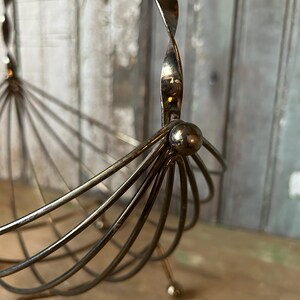 May include: A vintage silver-toned metal fruit basket with a unique design. The basket features a series of curved wires radiating from a central point, with a twisted handle. The base has small ball feet. The basket is on a wooden surface.