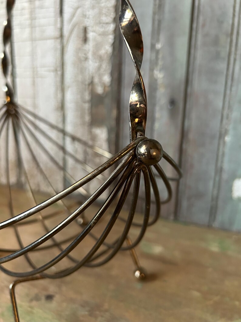 May include: A vintage silver-toned metal fruit basket with a unique design. The basket features a series of curved wires radiating from a central point, with a twisted handle. The base has small ball feet. The basket is on a wooden surface.