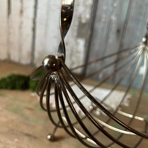 May include: A vintage metal fruit basket with a unique design. The basket features a spiraled handle and a series of curved metal rods forming the basket's body. The metal has a tarnished, aged appearance, adding to its vintage charm.