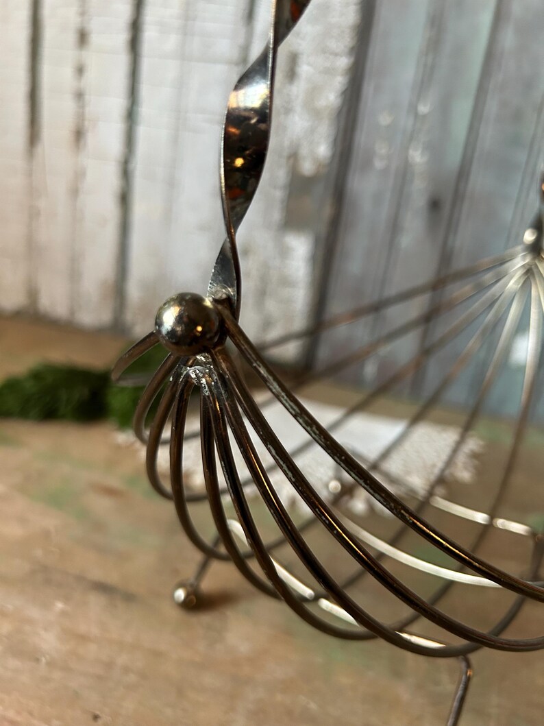 May include: A vintage metal fruit basket with a unique design. The basket features a spiraled handle and a series of curved metal rods forming the basket's body. The metal has a tarnished, aged appearance, adding to its vintage charm.