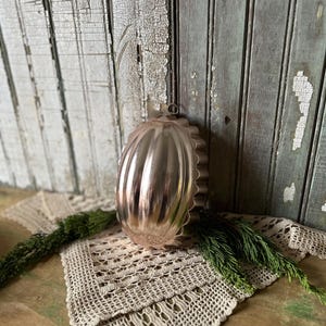 May include: A vintage, silver-colored metal mold with a fluted design, likely for baking. The mold has a loop for hanging. It rests on a cream-colored, crocheted doily with green sprigs. The background features weathered, painted wooden planks.