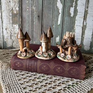 Może przedstawiać: Trzy ceramiczne pojemniki na wykałaczki w kształcie bajkowych domków. Każdy ma brązowy dach i beżowy korpus z dekoracyjnymi detalami. Pojemnik na wykałaczki po prawej stronie jest wypełniony wykałaczkami. Leżą na bordowej książce.