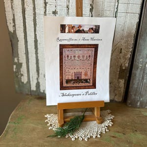 Op de afbeelding: Een ingelijste kruissteeksampler getiteld "Ragumuffin no. 1: Anne Harrison" door "Shakespeare's Peddler" wordt weergegeven op een kleine houten ezel. De sampler heeft een gedetailleerd ontwerp met een huis, bloemmotieven en tekst. Een takje groen en een kanten kleedje liggen eronder.