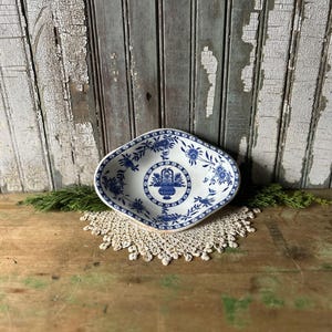 Op de afbeelding: Een wit en blauw decoratief schaaltje met een bloemmotief. Het schaaltje heeft een unieke vorm en ligt op een crèmekleurig kanten kleedje. De achtergrond toont verweerde houten planken.