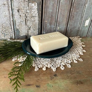 May include: A bar of soap with the text "ALL-ONE" embossed on top, resting on a dark green soap dish. The dish sits on a white lace doily, with a sprig of greenery to the left. The background is weathered wood.