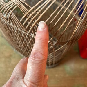 May include: Close-up of a woven basket made of light brown wicker. The basket has a rounded shape with a textured surface. A finger is pointing at the basket.