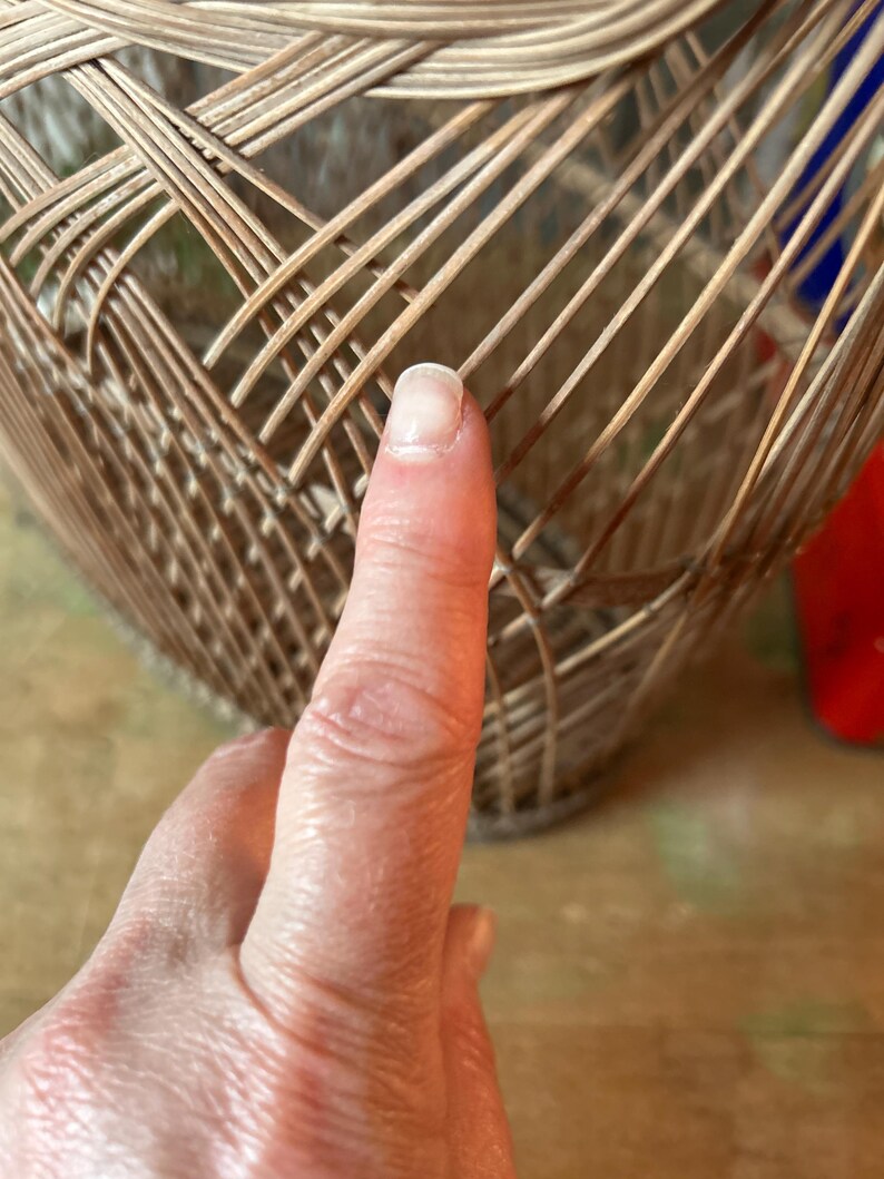 May include: Close-up of a woven basket made of light brown wicker. The basket has a rounded shape with a textured surface. A finger is pointing at the basket.