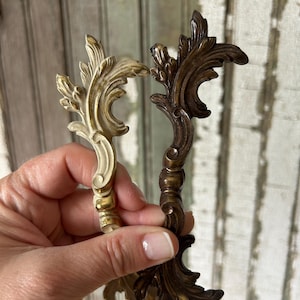 May include: Two decorative, ornate drawer pulls. One is a creamy off-white color with gold accents, and the other is a dark bronze. Both have a detailed, scrolling leaf design, perfect for adding a vintage touch to furniture.