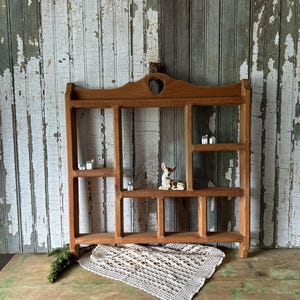 May include: Wooden wall shelf with multiple compartments, featuring a heart-shaped cutout at the top. The shelf is filled with miniature houses and a ceramic deer figurine. A crocheted doily and greenery are placed below the shelf.