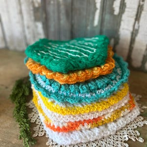 May include: A stack of colorful, handmade crochet washcloths. The washcloths are in shades of green, orange, turquoise, yellow, and white. The top washcloth is green with white stitching. The stack sits on a white lace doily.