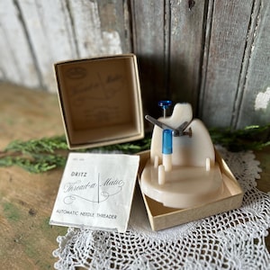 Puede incluir: Un enhebrador automático de agujas "Thread-a-Matic" de Dritz, en su caja original. El enhebrador es beige y azul, y la caja es de color canela. El producto se exhibe sobre un mantel individual de encaje blanco. El nombre del producto está impreso.