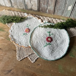 May include: Two white crocheted pot holders with embroidered floral designs. One has a green border and red flower, the other has an orange border and a pink flower. They rest on a beige lace doily.