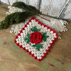 May include: A handmade crochet coaster featuring a central red rose with green leaves, surrounded by a cream-colored square with a red border. A sprig of green foliage and a lace doily are in the background.