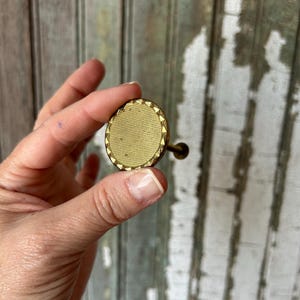 Peut inclure: Un bouton de meuble rond doré avec un centre texturé et une bordure géométrique décorative. Le bouton est tenu dans une main, mettant en valeur sa petite taille et son design complexe. Le bouton a une petite tige sombre.