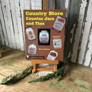 May include: A book titled "Country Store Counter Jars and Tins" is displayed on a small wooden easel. The cover features illustrations of vintage jars and tins, including "Gordon's Potato Chips" and "Chesty Potato Chips". The book is by Steve Batson.