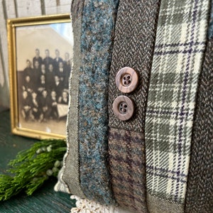 K&ouml;nnte beinhalten: Ein braunes und gr&uuml;nes Patchwork-Kissen mit zwei braunen Kn&ouml;pfen. Das Kissen ist aus verschiedenen Stoffen gefertigt, darunter karierter Stoff und Tweed.
