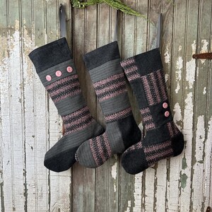 Può includere: Tre calze di Natale a righe grigie e rosa con bottoni rosa appese a una porta di legno invecchiata.