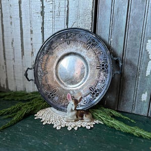 May include: A silver serving tray with an ornate design and two handles. The tray is sitting on a green surface with a small porcelain deer figurine and a lace doily.