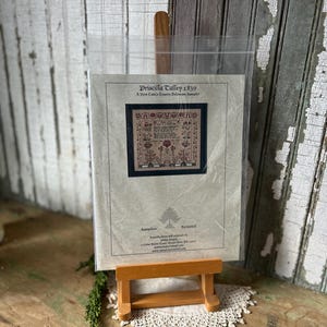 Peut inclure: Un échantillon de point de croix intitulé "Priscilla Talley 1839" dans une pochette en plastique transparent, exposé sur un petit chevalet en bois. L'échantillon présente un motif floral et textuel détaillé dans des tons de rouge, marron et vert.