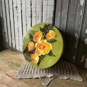 May include: A round, vintage green tin with a floral design featuring yellow roses and green leaves. The tin sits on a beige, crocheted doily. The background includes weathered, white and green wooden planks.