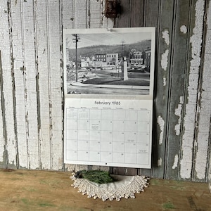 May include: A black and white calendar page for February 1985, featuring a town square photo. The calendar is hanging on a weathered, white and gray wooden wall. A small green and white decorative piece rests below.