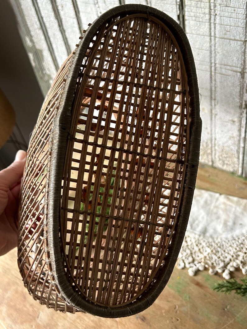 May include: A woven brown wicker basket with an oval shape. The basket has a dark brown trim around the top edge. The interior of the basket is visible through the woven pattern.