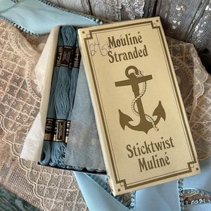 May include: A box of blue embroidery floss with a gold anchor and the text "Mouline Stranded Sticktwist Muline" on the front.