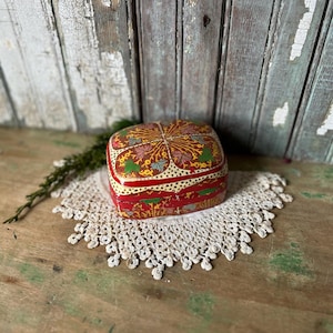 May include: A small, square, red and gold decorative box with floral patterns. The box sits on a white, crocheted doily. The background features weathered, painted wood.