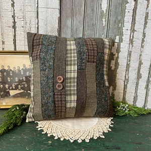 K&ouml;nnte beinhalten: Ein quadratisches Kissen mit Patchwork-Muster aus verschiedenen Gr&uuml;nt&ouml;nen, Braun- und Blaut&ouml;nen. Das Kissen hat zwei braune Kn&ouml;pfe in der Mitte.