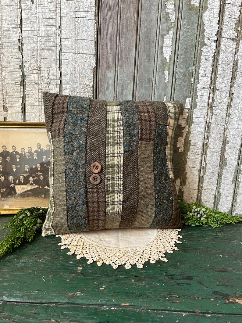 K&ouml;nnte beinhalten: Ein quadratisches Kissen mit Patchwork-Muster aus verschiedenen Gr&uuml;nt&ouml;nen, Braun- und Blaut&ouml;nen. Das Kissen hat zwei braune Kn&ouml;pfe in der Mitte.