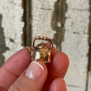 May include: A miniature gold-coloured teapot charm with a curved handle and spout. The small teapot has a rounded body and a tiny lid. The charm is held in a person's hand against a blurred, weathered wood background.