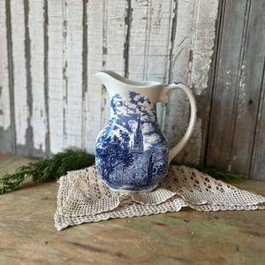 May include: A white ceramic pitcher featuring a blue transferware pattern depicting a church with a tall steeple, trees, and clouds. The pitcher has a curved handle, a pouring spout, and a rounded body with a detailed landscape scene in blue ink.