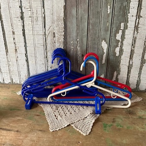 May include: A collection of blue, red, and white plastic clothes hangers. The hangers are arranged on a beige crocheted doily, with a weathered wooden background. The hangers have clips and hooks for hanging clothes.