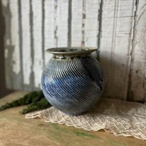 May include: A blue and white ceramic vase with a textured, ridged surface. The vase has a rounded shape with a slightly flared rim. The color transitions from a dark blue at the top to a lighter blue towards the bottom. The vase is displayed on a wooden surface.