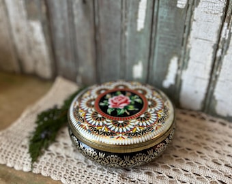 May include: A round, vintage metal tin with a floral design on the lid. The lid features a central rose surrounded by a decorative pattern in red, yellow, and white. The tin's sides are black with a gold trim.